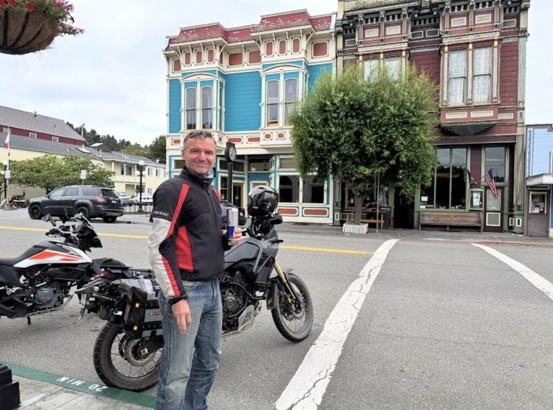 Man in motorcycle riding jacket standing in front of two adventure bikes and colorful Victorian-era storefronts on a small-town main street