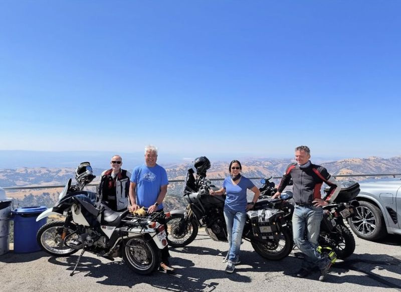 Four riders posing with adventure motorcycles at a hilltop overlook, rolling brown hills and blue sky stretching to the horizon